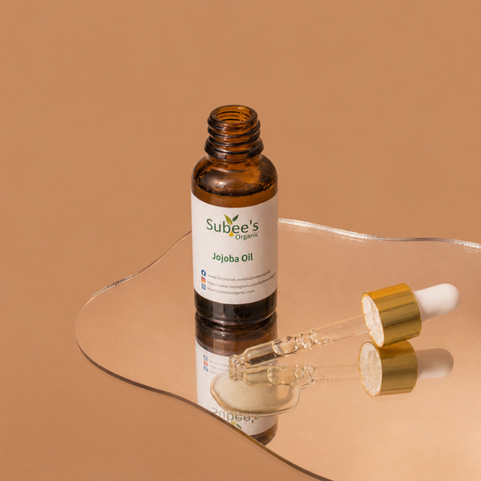 A bottle of Subee's Organics jojoba Oil with a glass dropper and gold cap resting on a clear, wavy glass tray against a solid warm beige background.