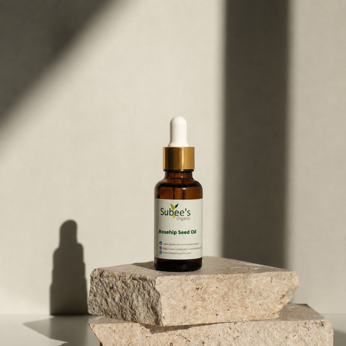 A small amber glass dropper bottle labeled 'Subbee's Organics Rosehip Seed Oil' sits on stacked stone blocks against a warm beige wall with strong sunlight and shadows.