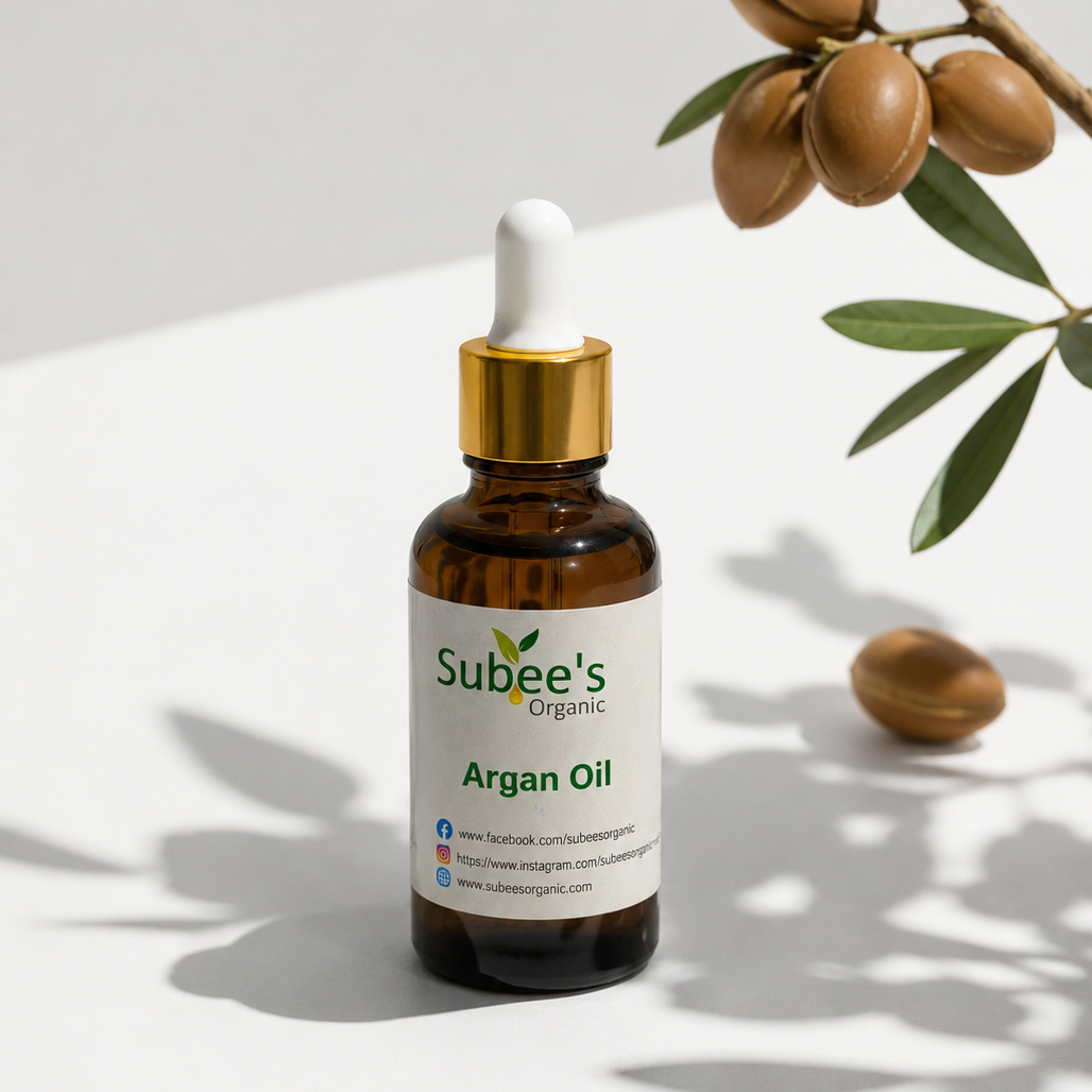 A bottle of Subee's Organic Argan Oil with a white dropper cap and gold collar, set on a white surface with argan tree leaves and nuts in the background.